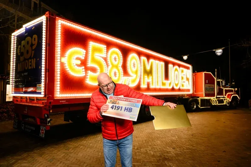 De verlichte prijzentruck van de Postcode Loterij bij avond, met een man die een prijscheque omhoog houdt.