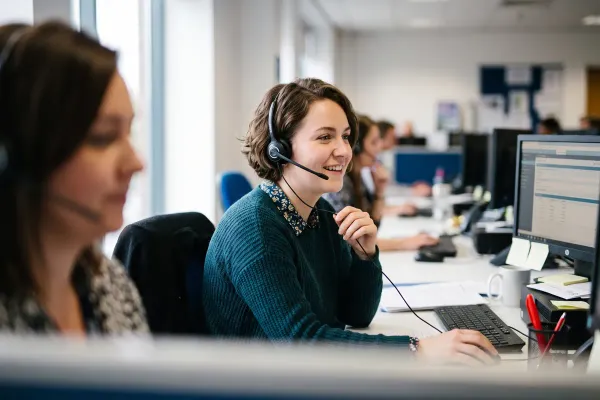 Een klantcontactmedewerker met headset helpt lachend een klant aan de telefoon, met haar scherm naast zich.